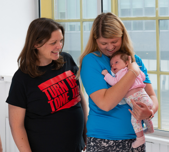 A woman wearing a blue top holding a young baby, a woman with a black top stands beside her